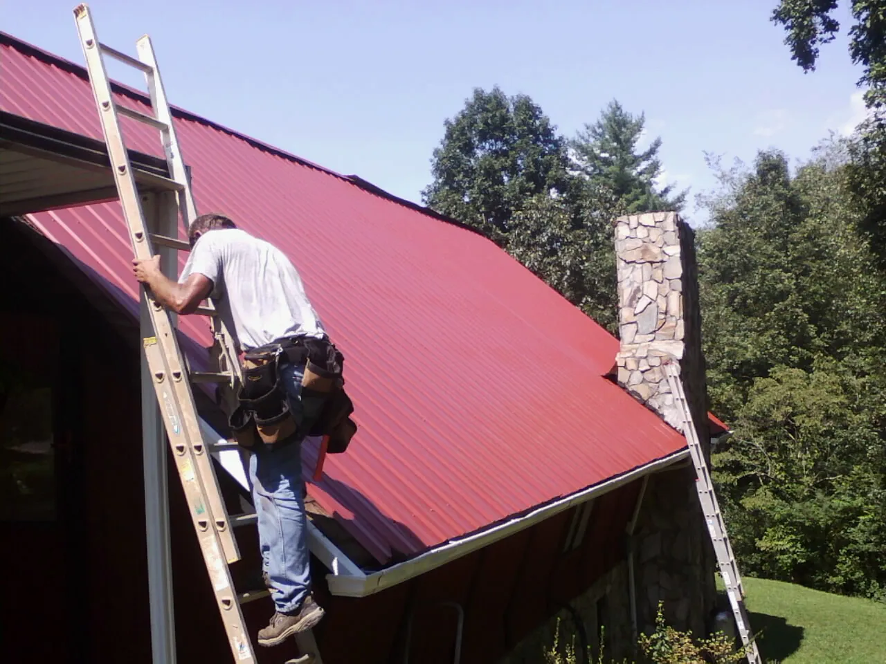 Roofer on a ladder beside a red metal roof and stone chimney — Soffit & Fascia Repair by Roofing Brothers in Indian Wells